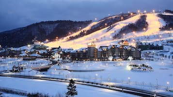 A snow-covered resort with multiple buildings, a ski slope, and a well-lit parking area.