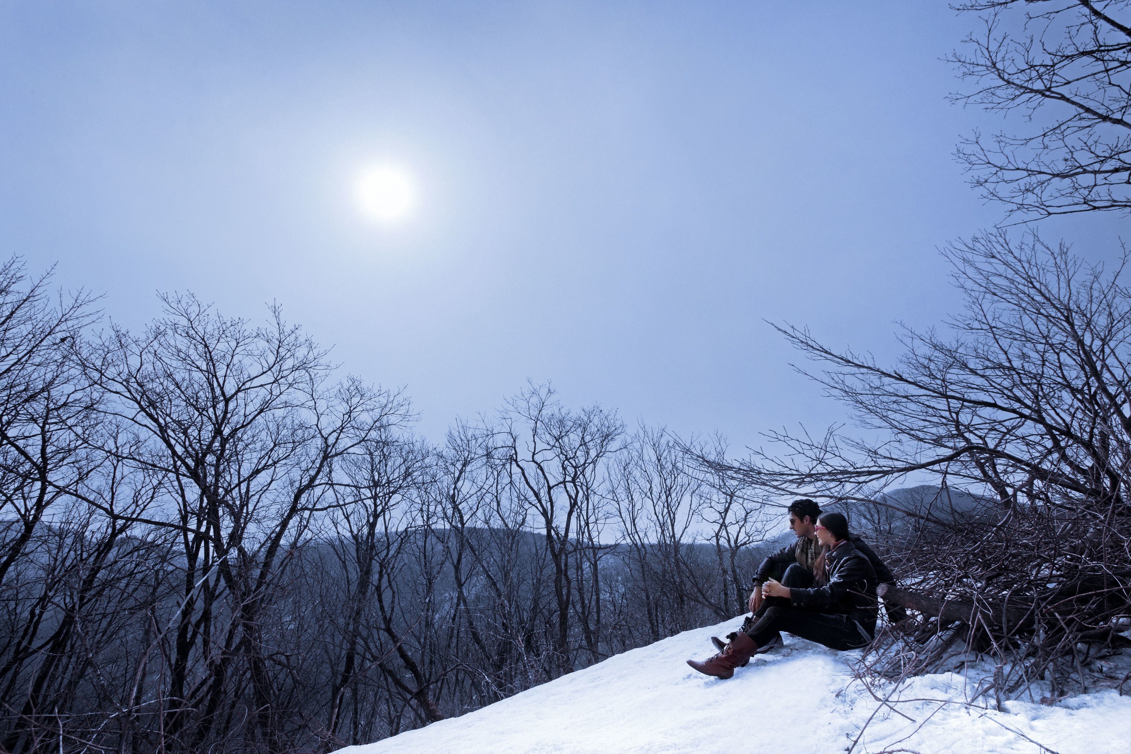 Two people sitting on a snow-covered slope with a clear sky and mountains in the background.