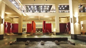 Un vestíbulo amplio con una fuente central, zonas de estar rojas y un balcón en el segundo piso con barandas de cristal.