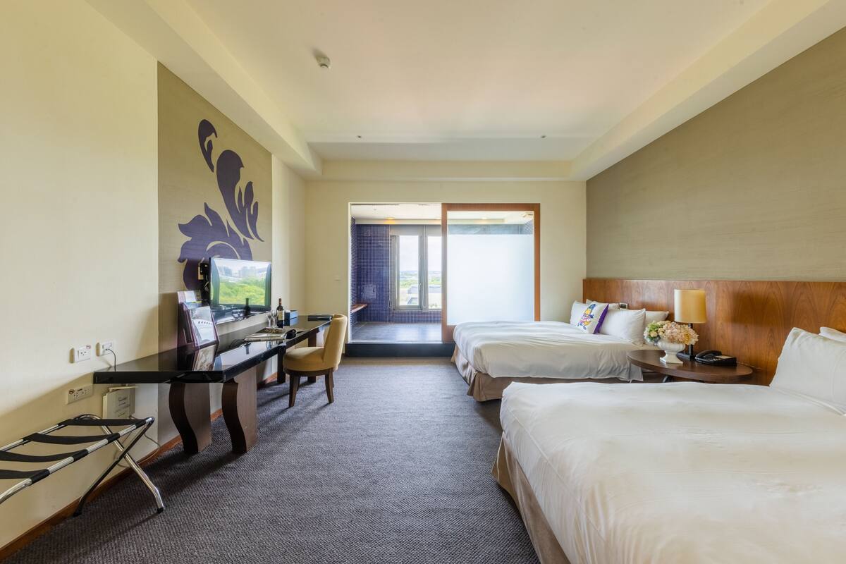 A hotel room with two beds, a desk with a chair, a TV, and a view of the outdoors through a sliding door.