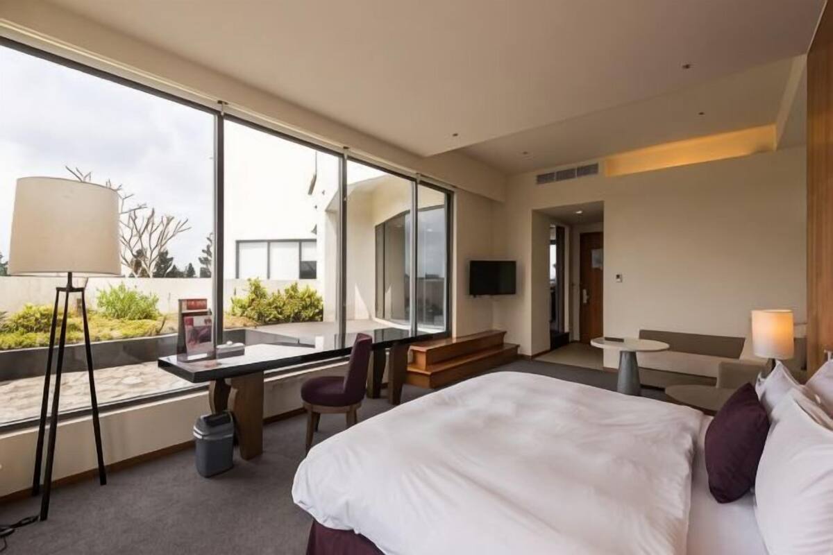 A modern hotel room with a large bed, a desk, a chair, a TV, and a view of the outdoors.