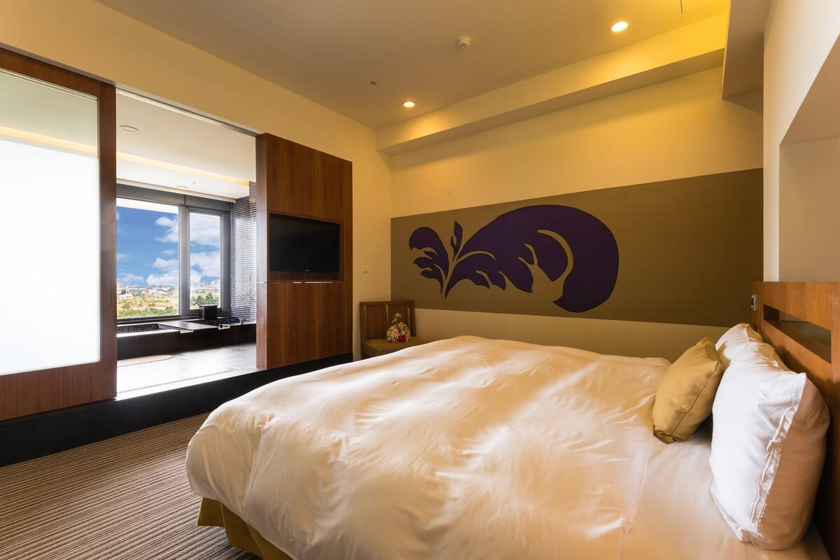 A modern hotel room with a large bed, a flat-screen TV, a window with a city view, and a decorative wall piece.