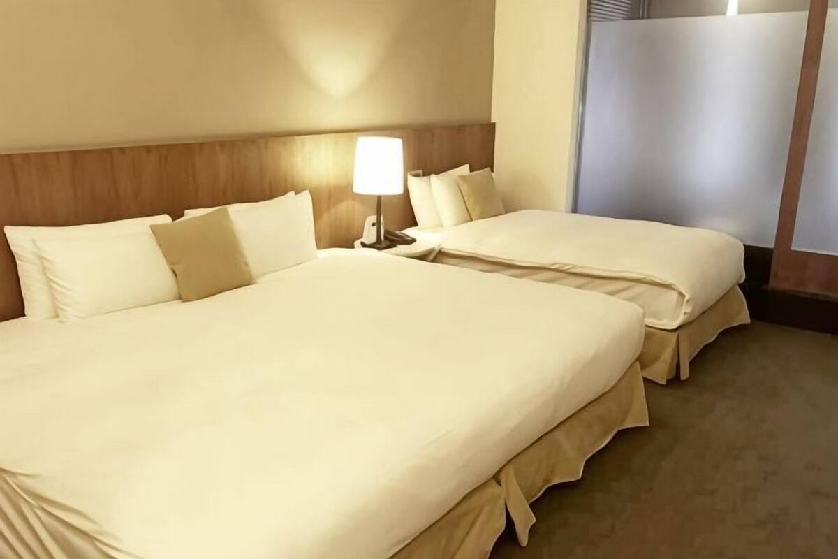 A hotel room with two beds, a nightstand with a lamp, and a window with a view.