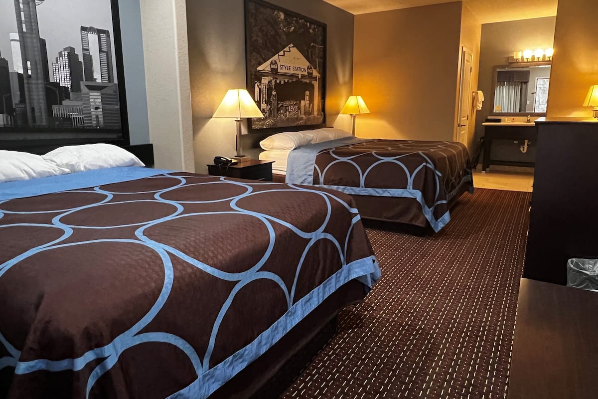 A hotel room with two beds, a cityscape painting, a desk with a lamp, and a mirror.