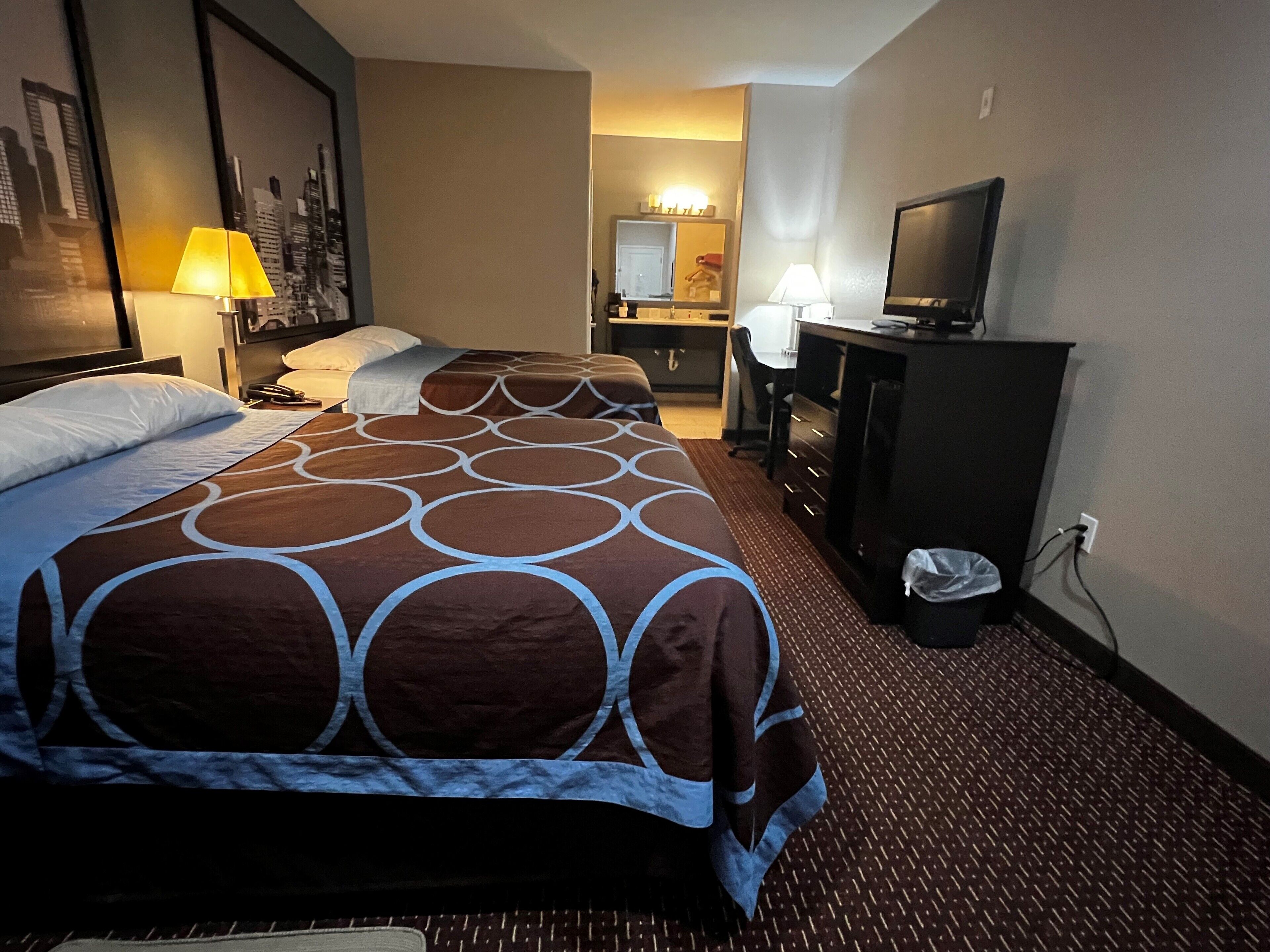 A hotel room with two beds, a desk, a television, and a bathroom in the background.