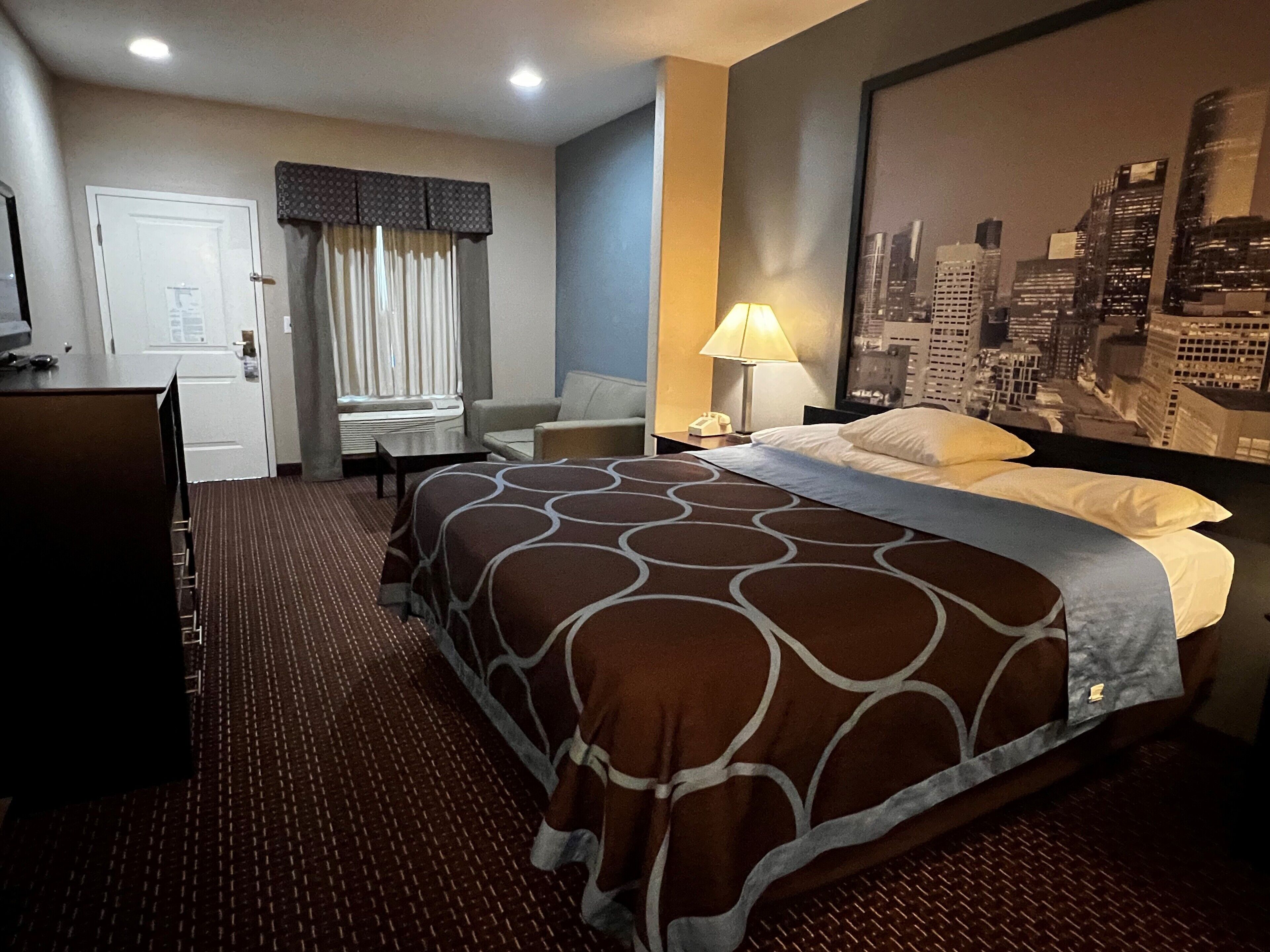 A hotel room with a bed, a desk, a chair, a lamp, and a cityscape view.