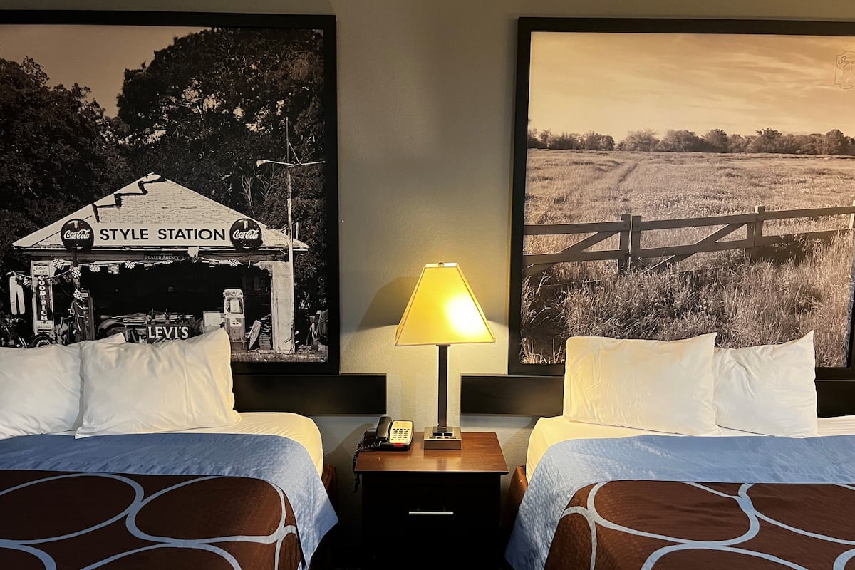 A hotel room with two beds, a nightstand with a lamp, and two large framed pictures on the wall.