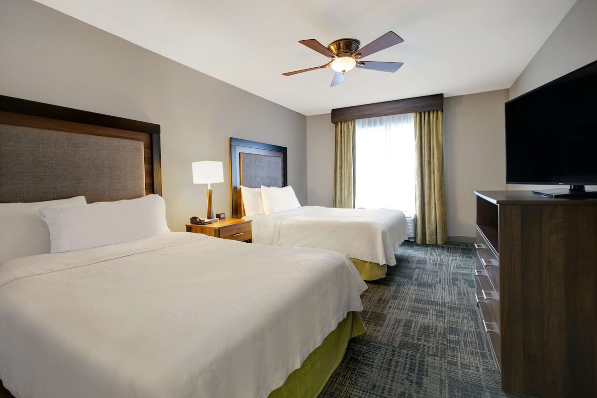 A hotel room with two beds, a ceiling fan, a flat-screen TV, and a large window with curtains.