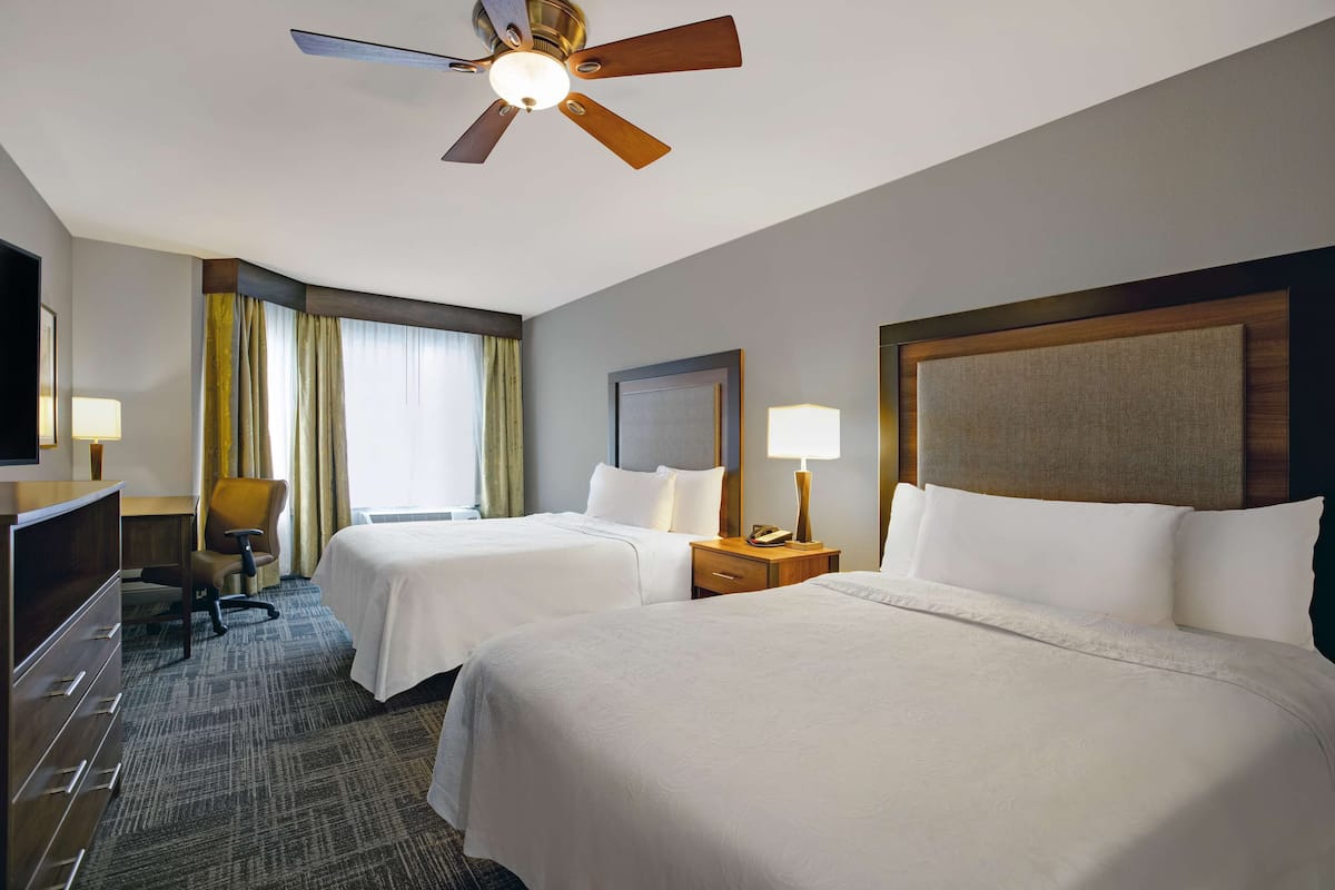 A hotel room with two beds, a desk, a chair, a ceiling fan, and a TV.