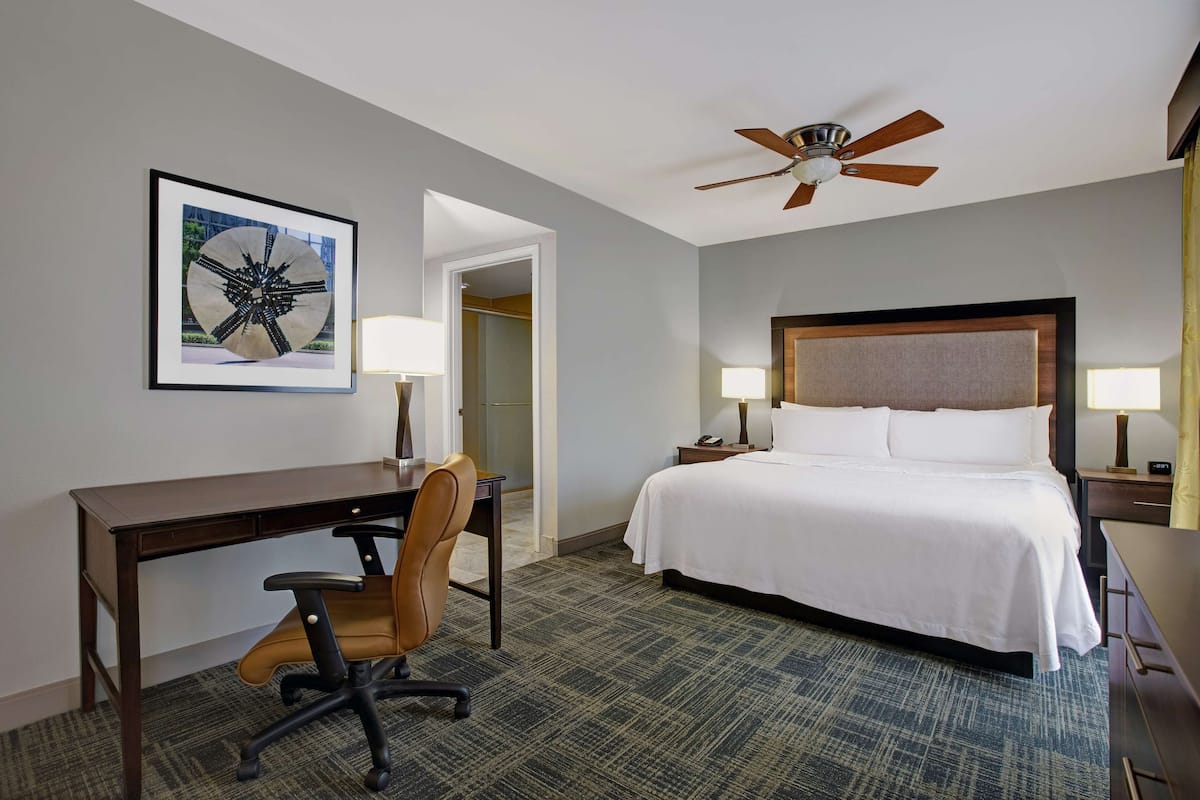 A hotel room with a bed, desk, chair, two bedside tables with lamps, a ceiling fan, and a framed picture on the wall.
