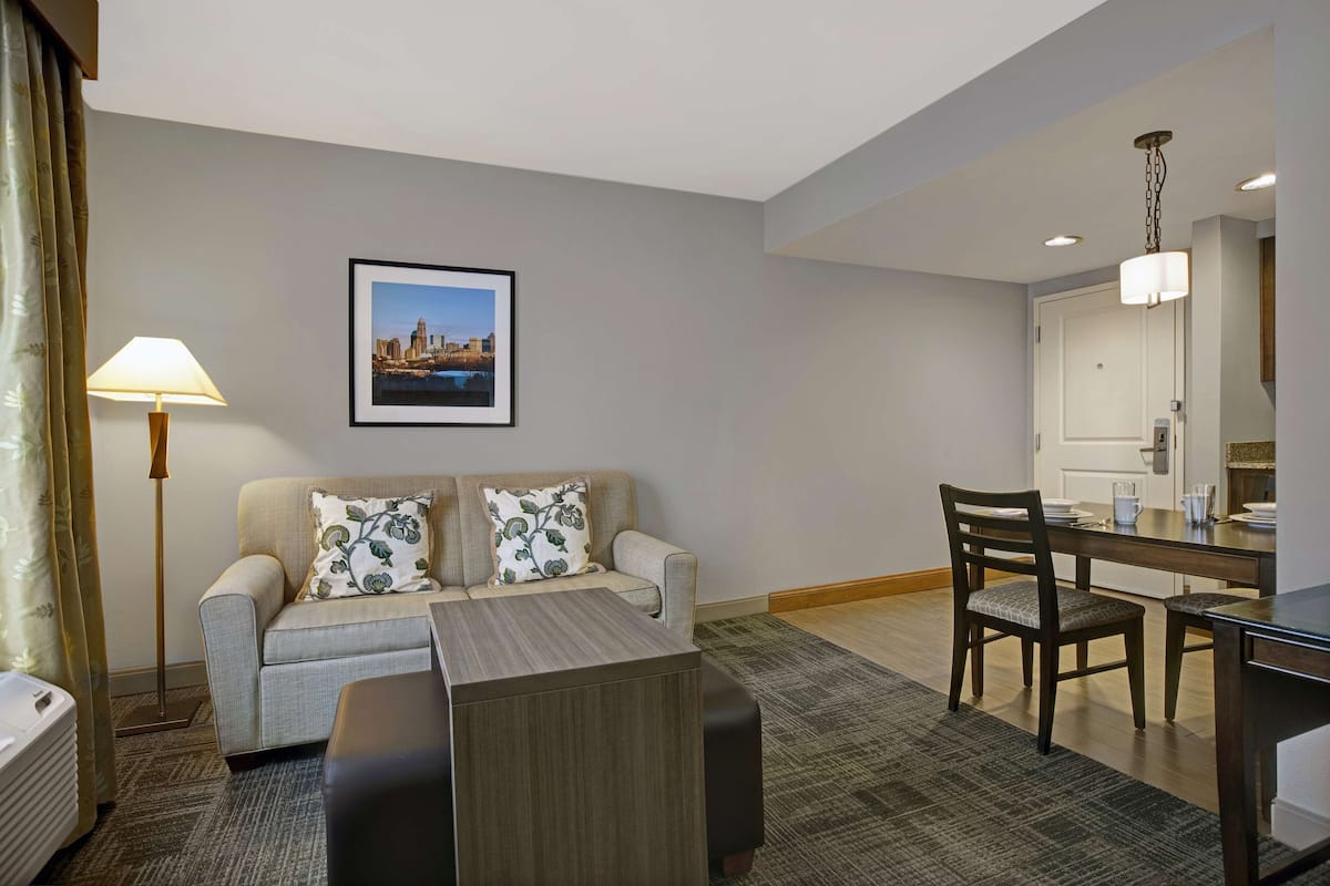 A hotel room with a sofa, a coffee table, a dining table with chairs, a lamp, and a framed picture on the wall.
