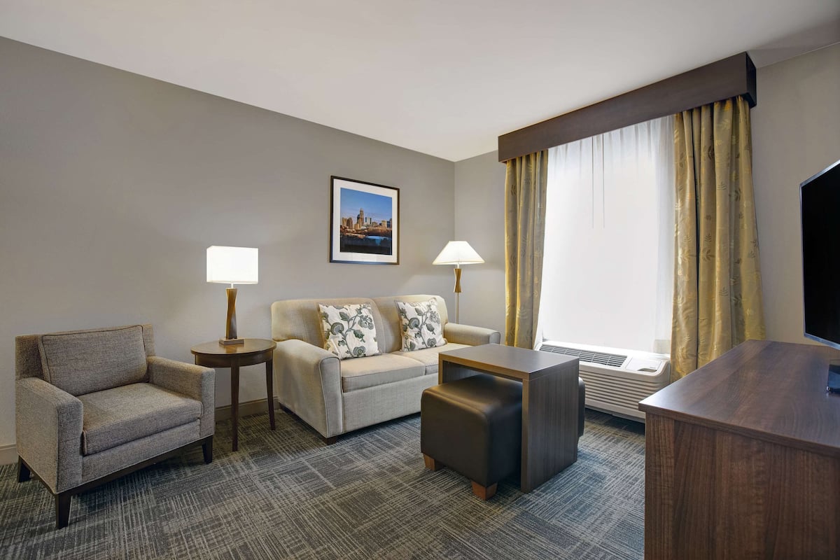 A hotel room with a sofa, armchair, and a small table. There is a picture on the wall and a window with curtains.