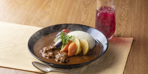 A bowl of stew with vegetables and rice, accompanied by a glass of iced drink on a wooden table.
