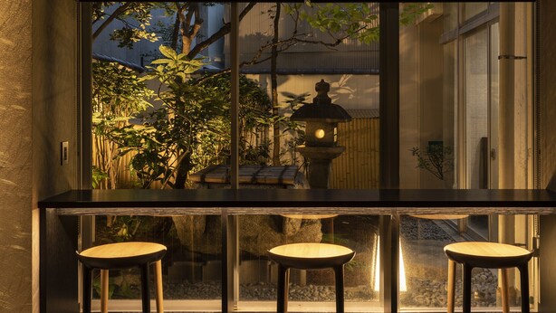 En bar med tre barstole, en stenen lanterne og udsigt til en have med bambus og en traditionel japansk bygning.