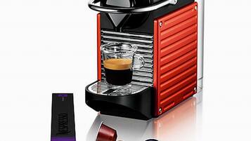 Eine Nespresso-Kaffeemaschine mit rotem und schwarzem Design, eine Glaskaraffe und zwei Kapseln.
