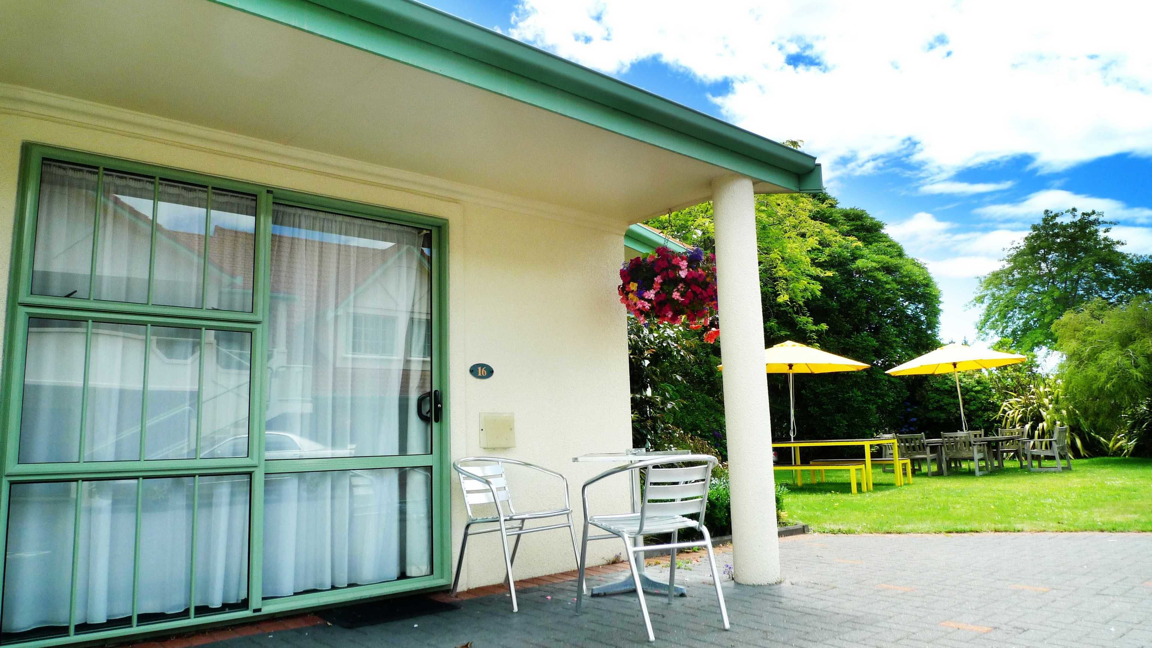 A building with a green-framed window, a door, and a number '16' sign. There are two metal chairs and a yellow umbrella outside.