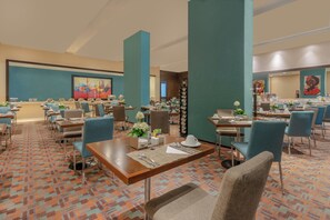 A hotel dining area with multiple tables and chairs, a patterned carpet, and teal-colored walls. - Hotel Estelar En Alto Prado (Barranquilla)
