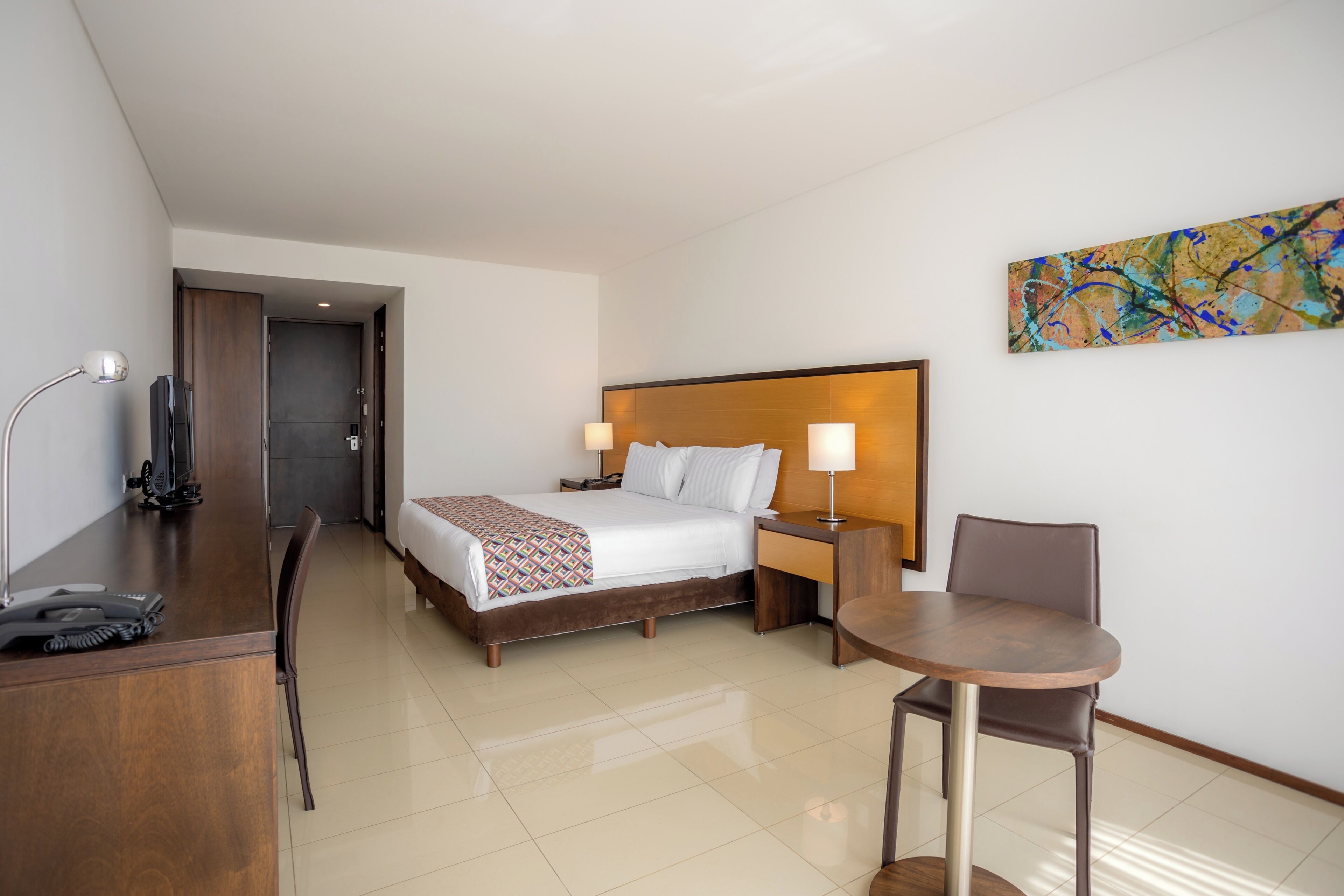 Habitación de hotel con cama, mesita de noche, lámpara, escritorio, silla y un cuadro en la pared.