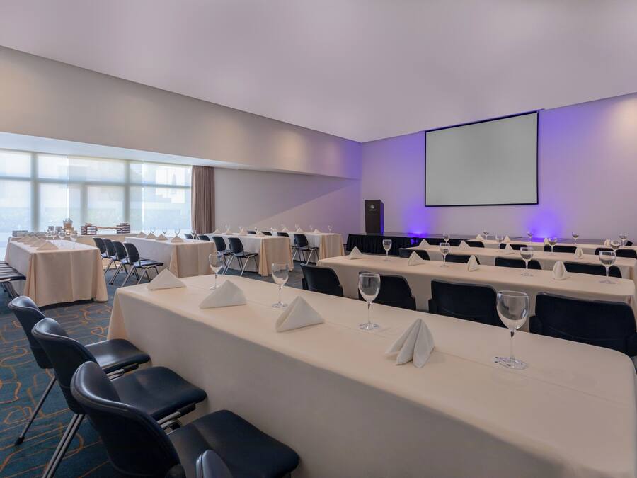 Un salón de conferencias con mesas dispuestas para reuniones, una pantalla de proyector y un ventanal grande.