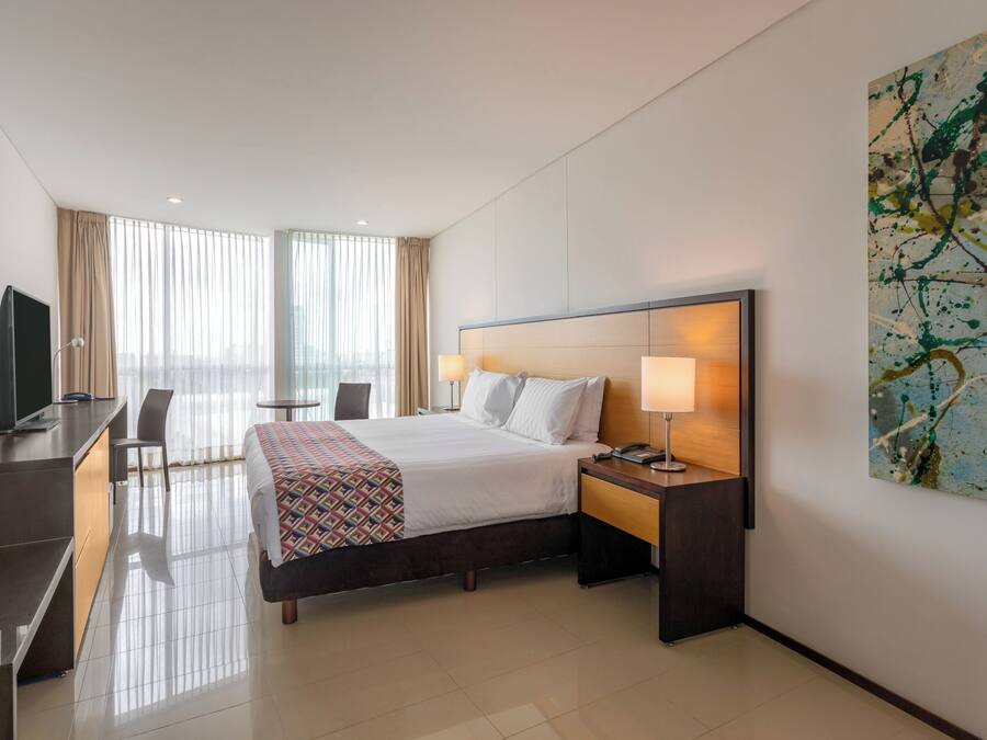 Habitación de hotel con cama, mesita de noche, lámpara, televisor, escritorio, silla y un cuadro abstracto grande en la pared.
