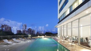 Una piscina sul tetto con lettini e tavoli, uno skyline cittadino sullo sfondo e un edificio moderno con ampie vetrate.