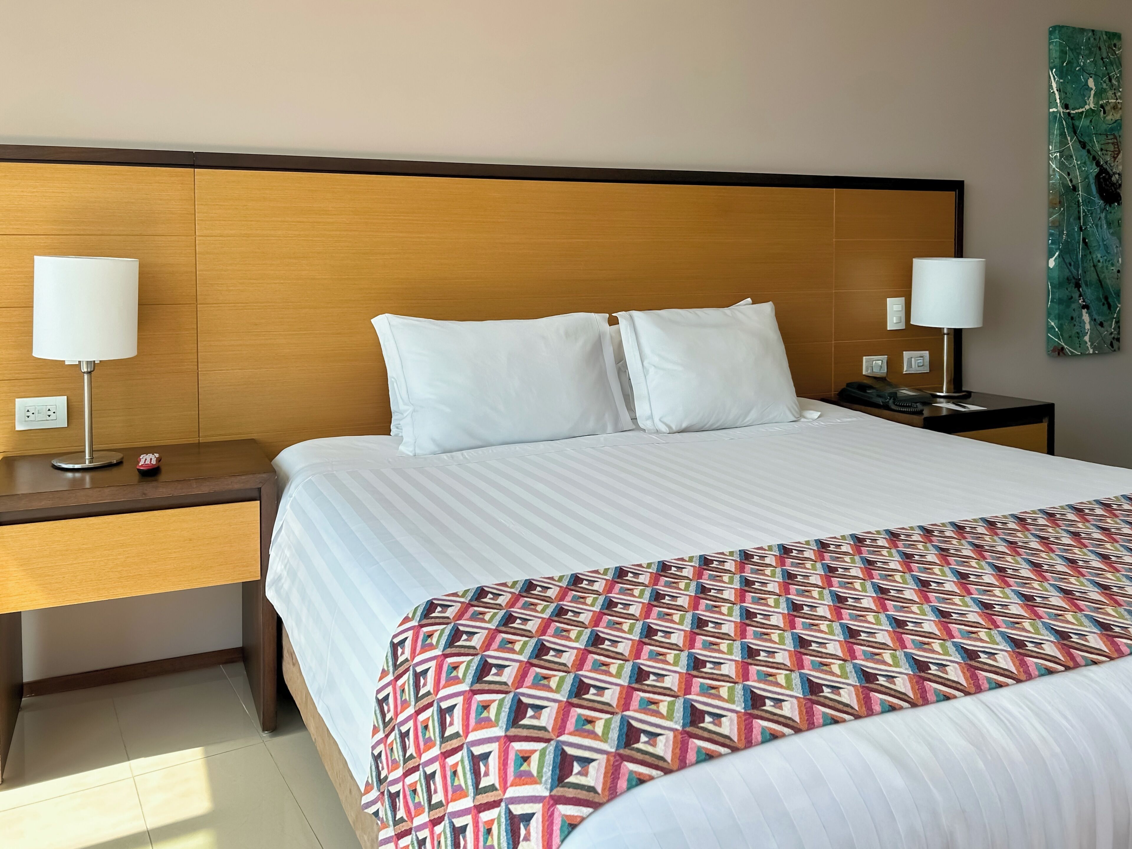 Habitación de hotel con una cama grande, dos mesitas de noche, una lámpara en cada una, un teléfono y una obra de arte en la pared.