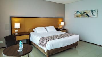 Habitación de hotel con cama, dos mesitas de noche con lámparas, una silla, una pequeña mesa redonda con una botella de agua y un cuadro en la pared.