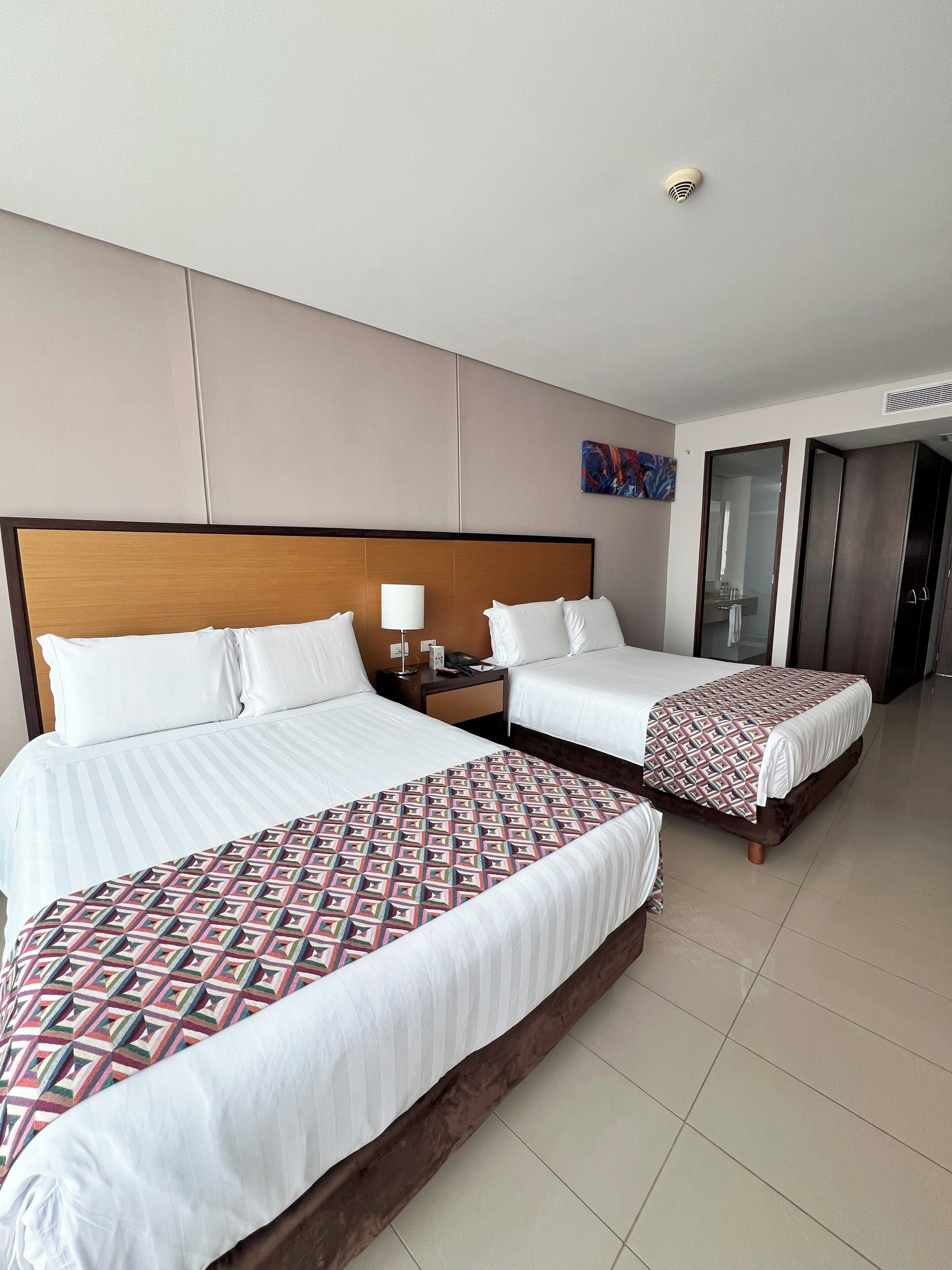A hotel room with two beds, a nightstand, a lamp, a painting on the wall, and a door leading to another room.