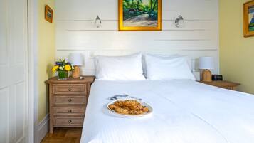 Uma cama bem arrumada com lençóis brancos, uma mesa de cabeceira com abajur, um pequeno vaso com flores e um prato de biscoitos.