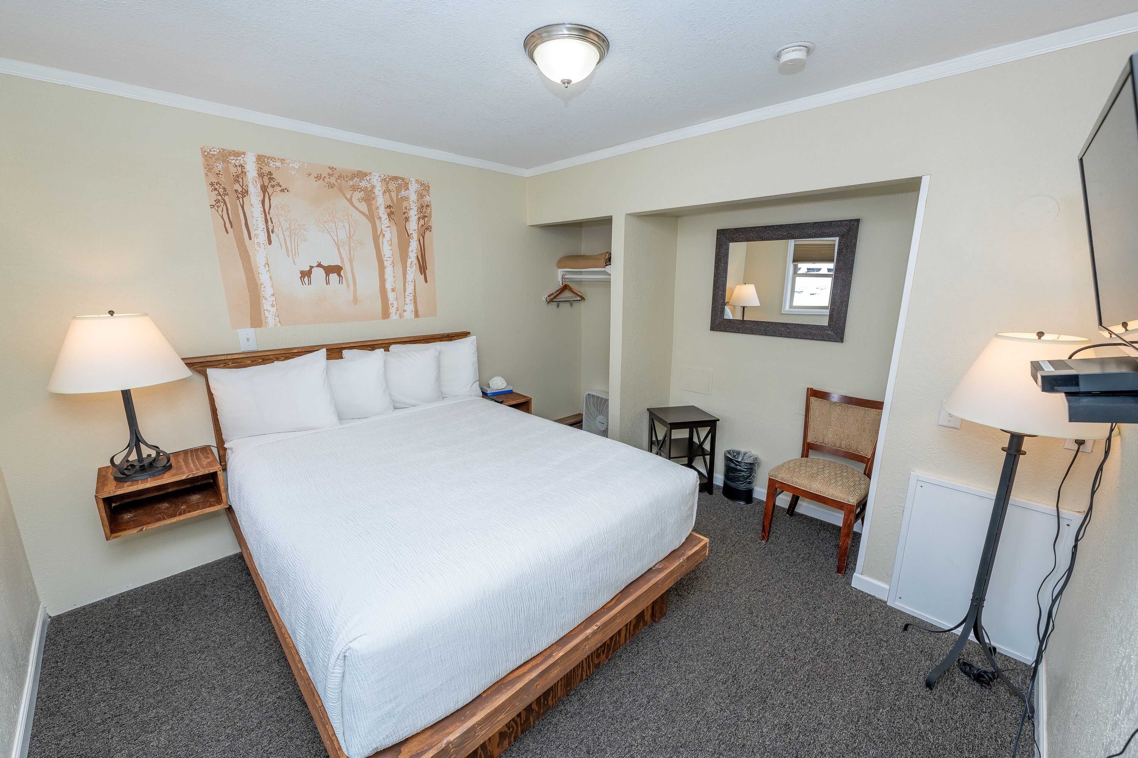 A hotel room with a large bed, bedside tables with lamps, a chair, a small table, a mirror, and a wall art piece.