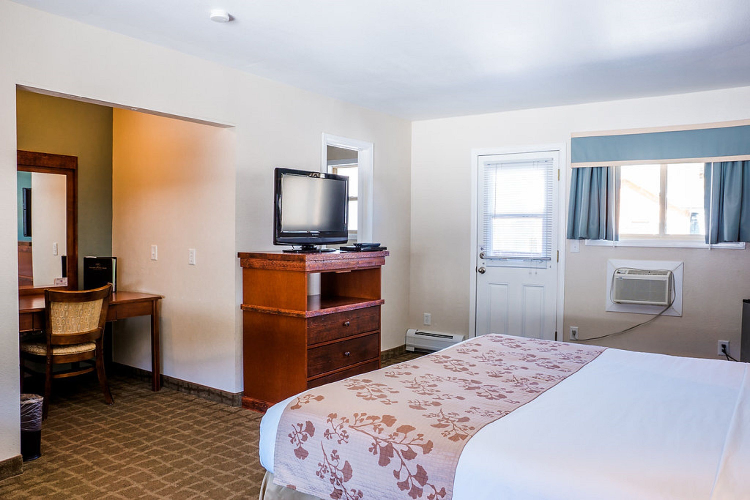 A hotel room with a bed, a desk with a chair, a television, a wardrobe, a door, and a window with curtains.