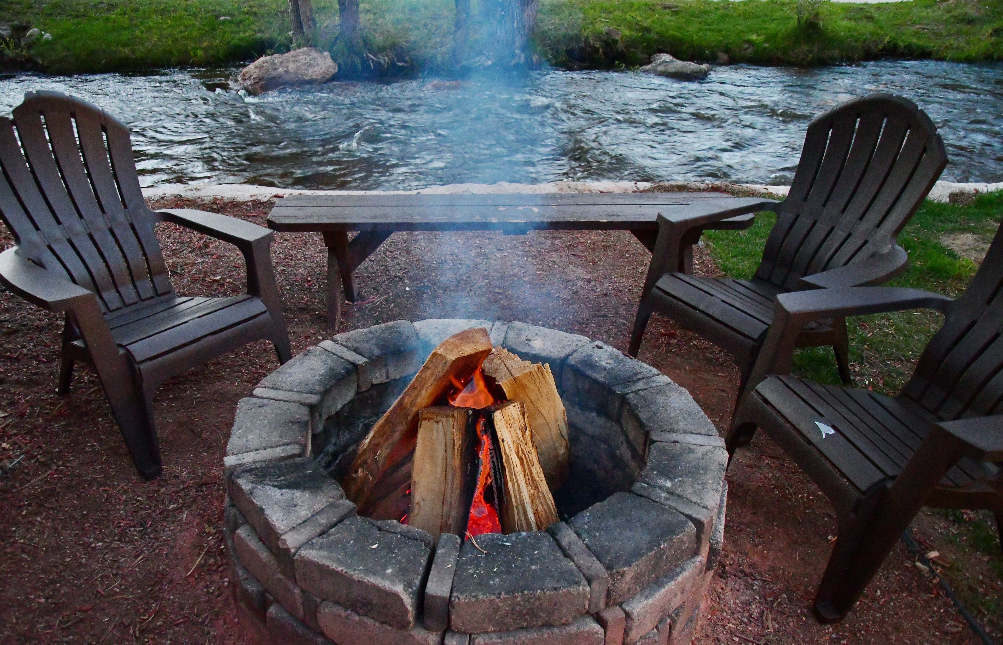 Ein Lagerfeuer mit zwei Adirondack-Stühlen und einem Tisch in einer natürlichen Umgebung, im Hintergrund fließt ein Fluss.