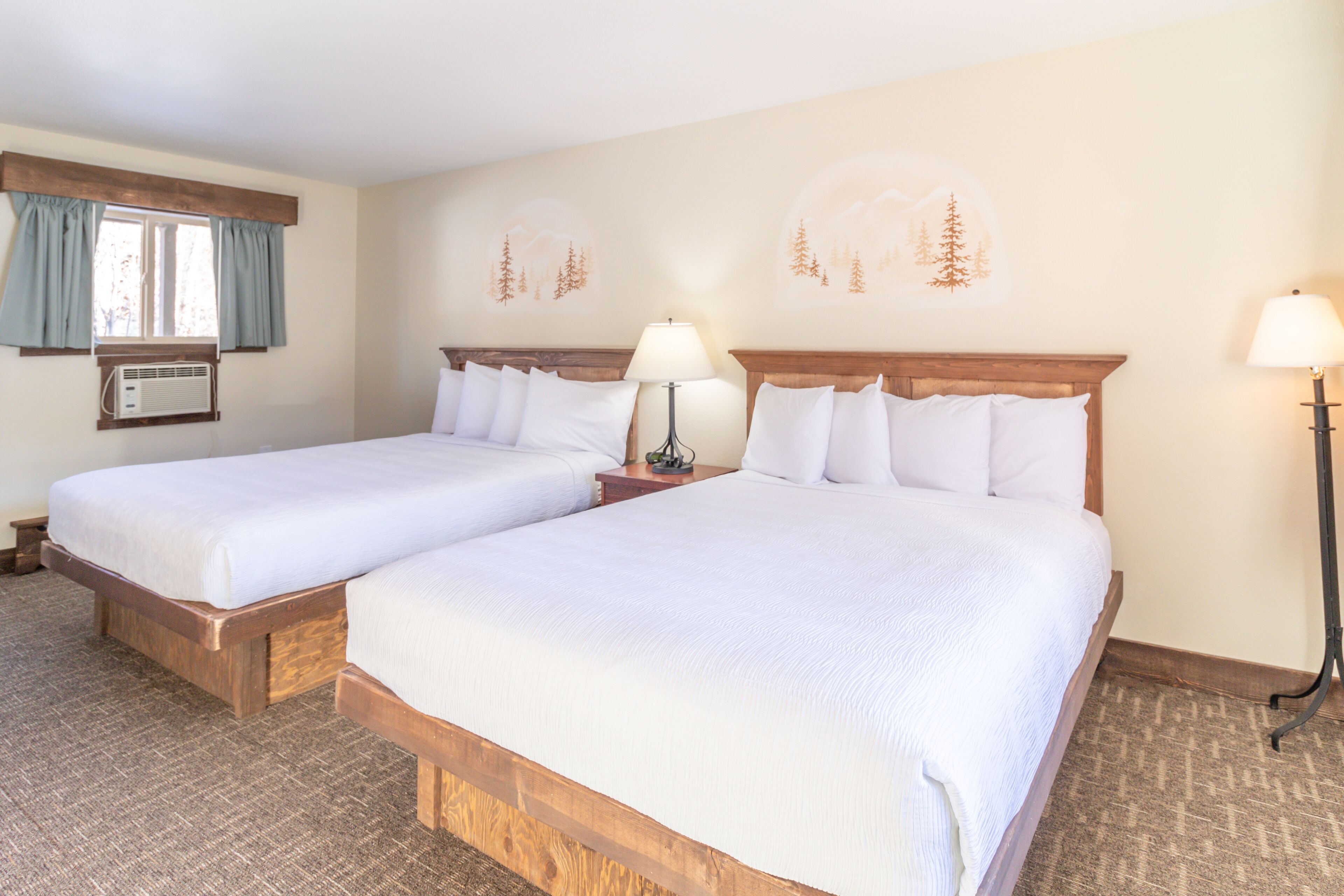Une chambre d’hôtel avec deux lits simples, une fenêtre avec des rideaux, un appareil de climatisation et des œuvres d’art murales représentant des arbres.