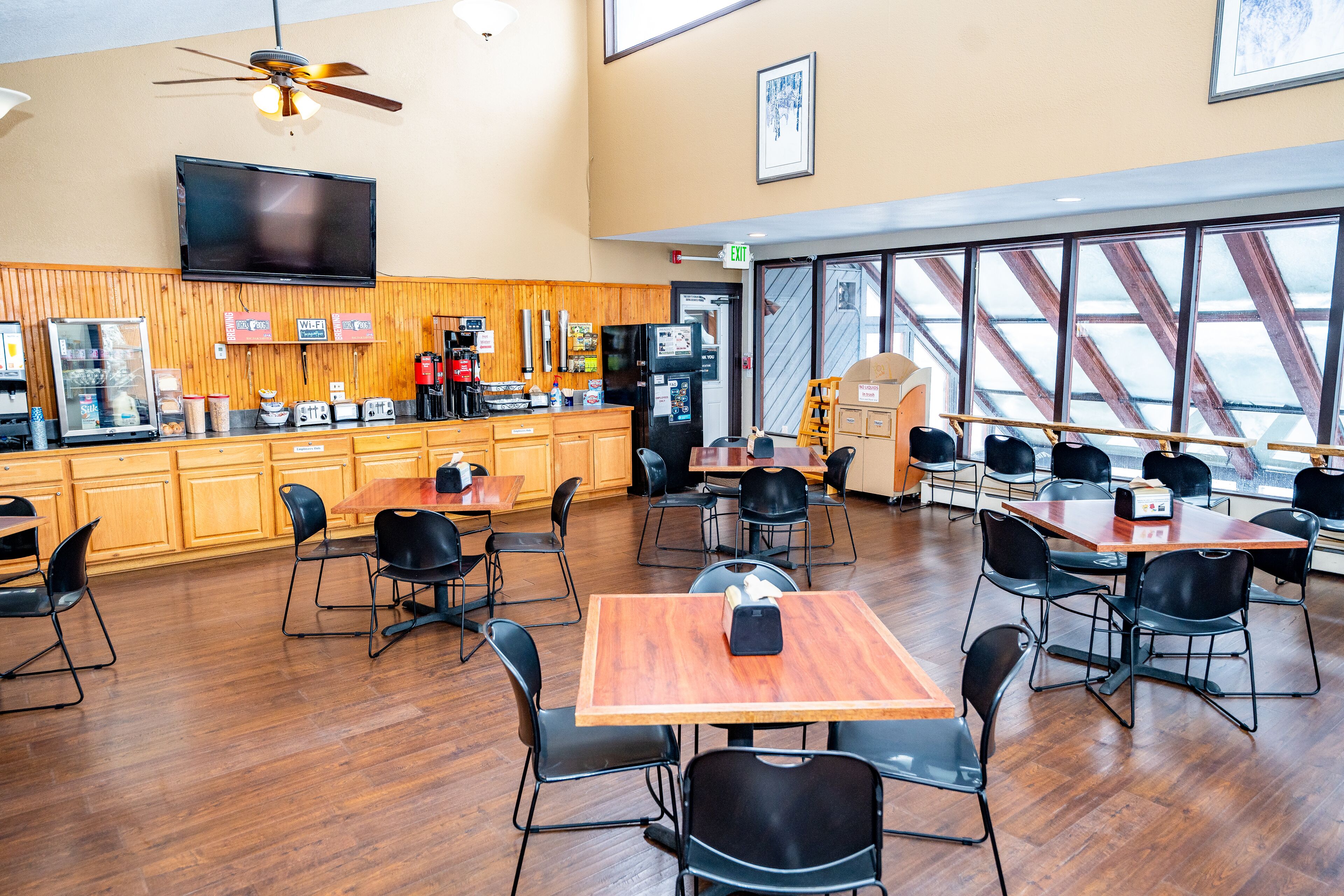 A spacious indoor dining area with wooden tables and black chairs, a kitchen counter with appliances, and large windows with a view.