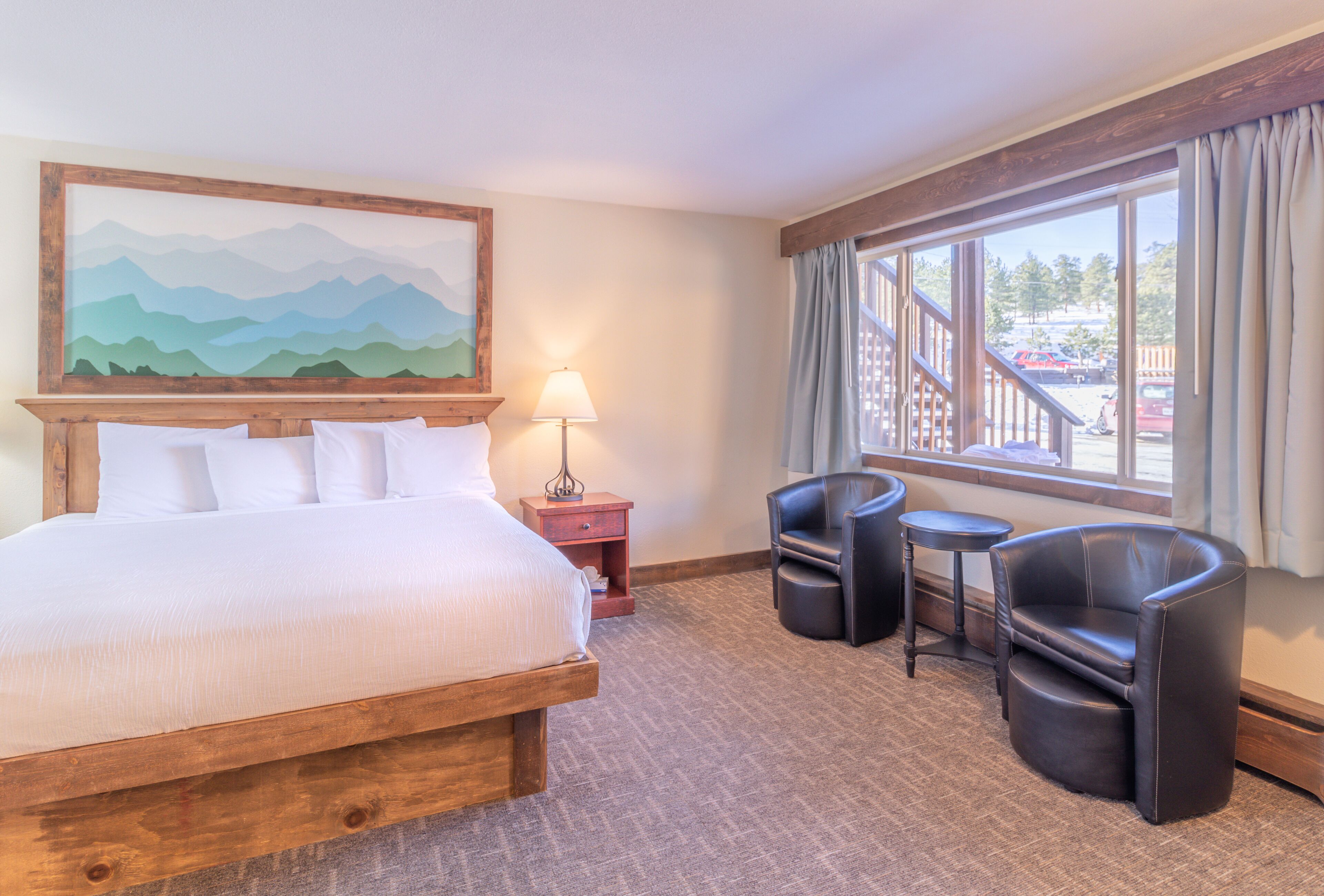 A hotel room with a large bed, a desk with two chairs, a lamp, and a window with a view of a parking area.