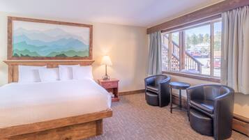 A hotel room with a large bed, a desk with two chairs, a lamp, and a window with a view of a parking area.