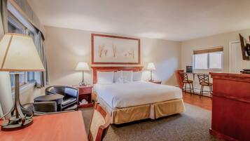 A hotel room with a large bed, two bedside tables with lamps, a desk with two chairs, a red cabinet, and a window with blinds.