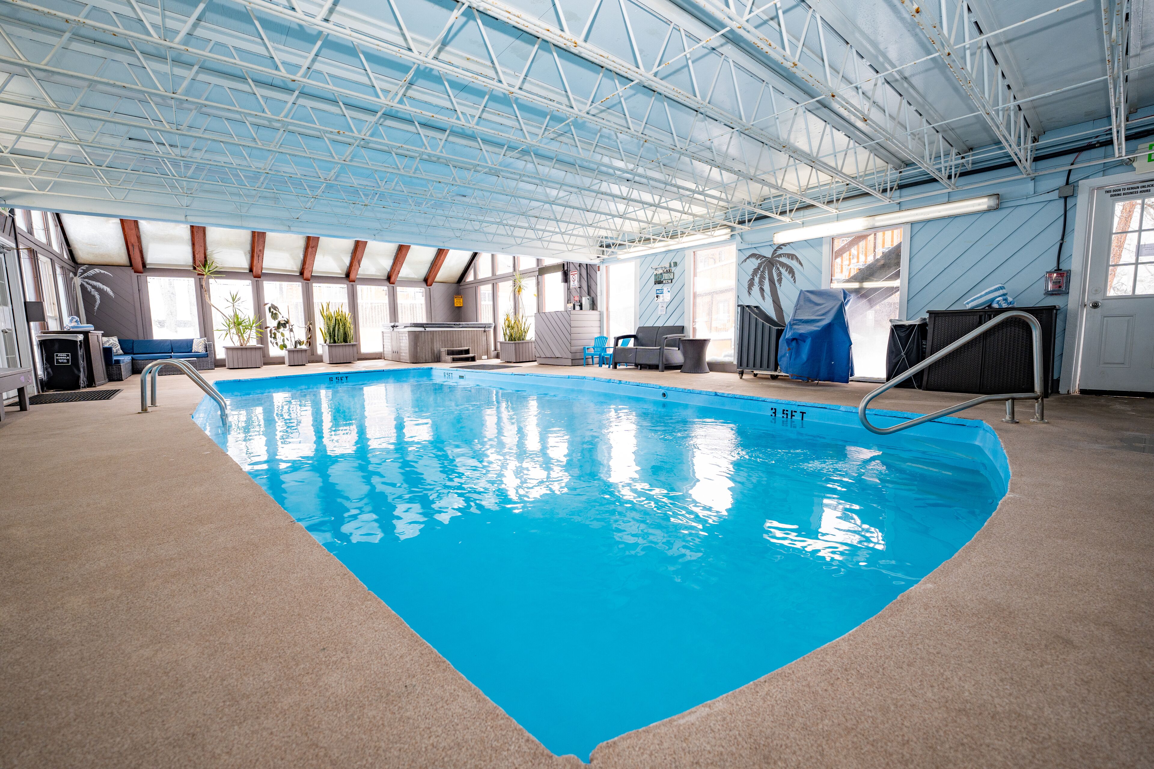 Une piscine intérieure à l’eau d’un bleu cristallin, entourée d’un sol de couleur beige et surmontée d’un plafond haut avec des poutres métalliques.
