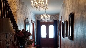 Un couloir agrémenté d’un lustre, d’un parquet en bois, d’un tapis et d’un escalier dont les murs sont recouverts d’un papier peint à motifs.