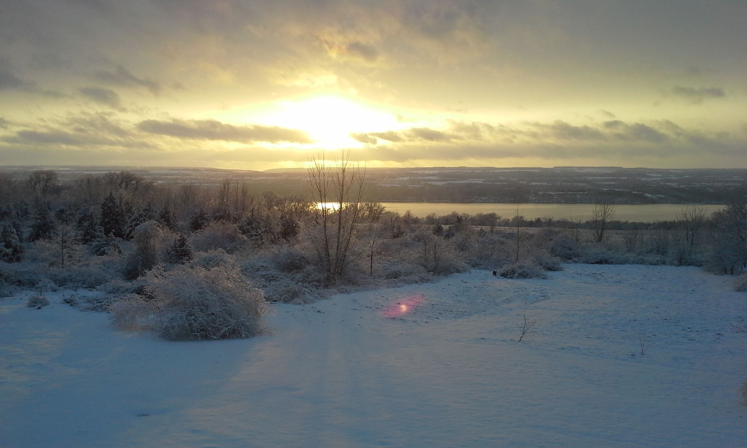 A snowy landscape with a sunset over a body of water and distant hills.