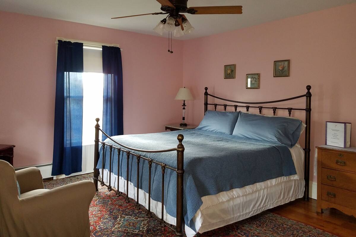A bedroom with a bed, a nightstand, a chair, a ceiling fan, and a window with curtains.