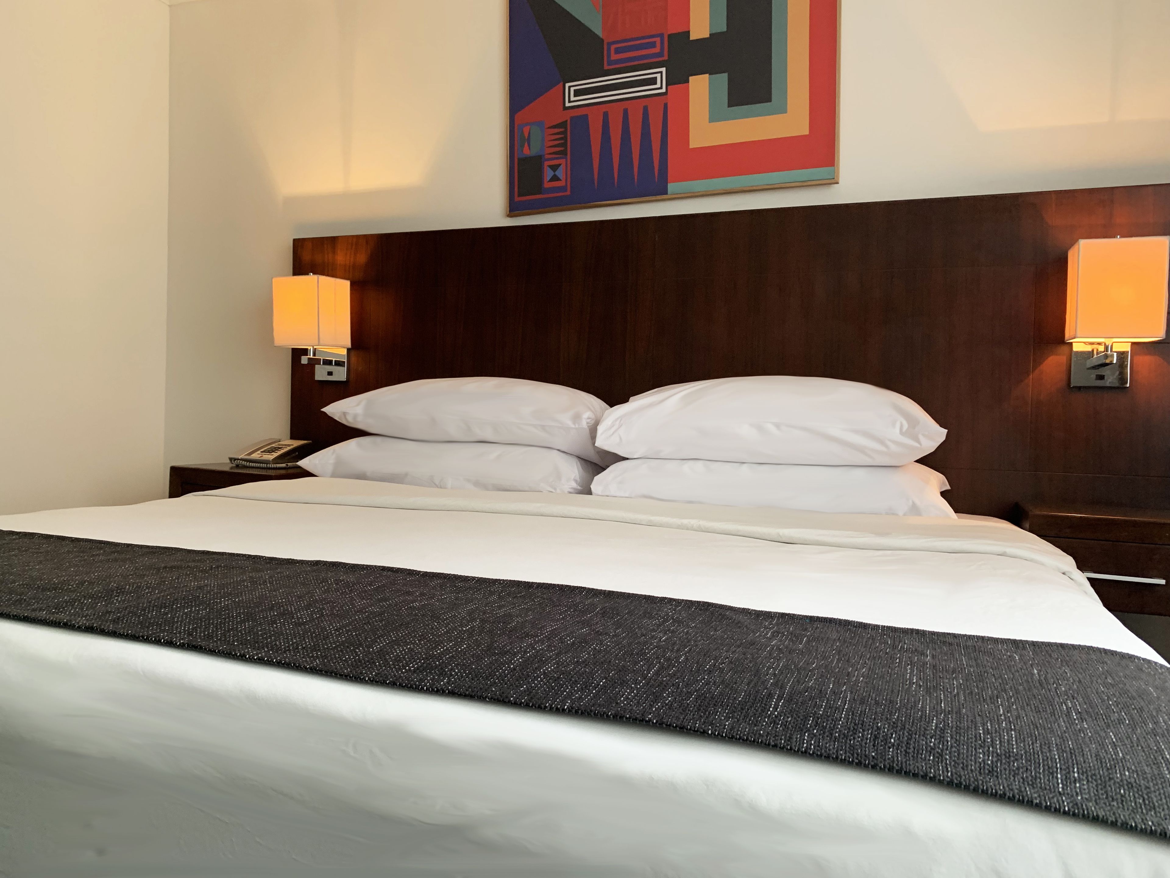 A hotel room with a large bed, two bedside lamps, a telephone, and a colorful abstract painting on the wall.