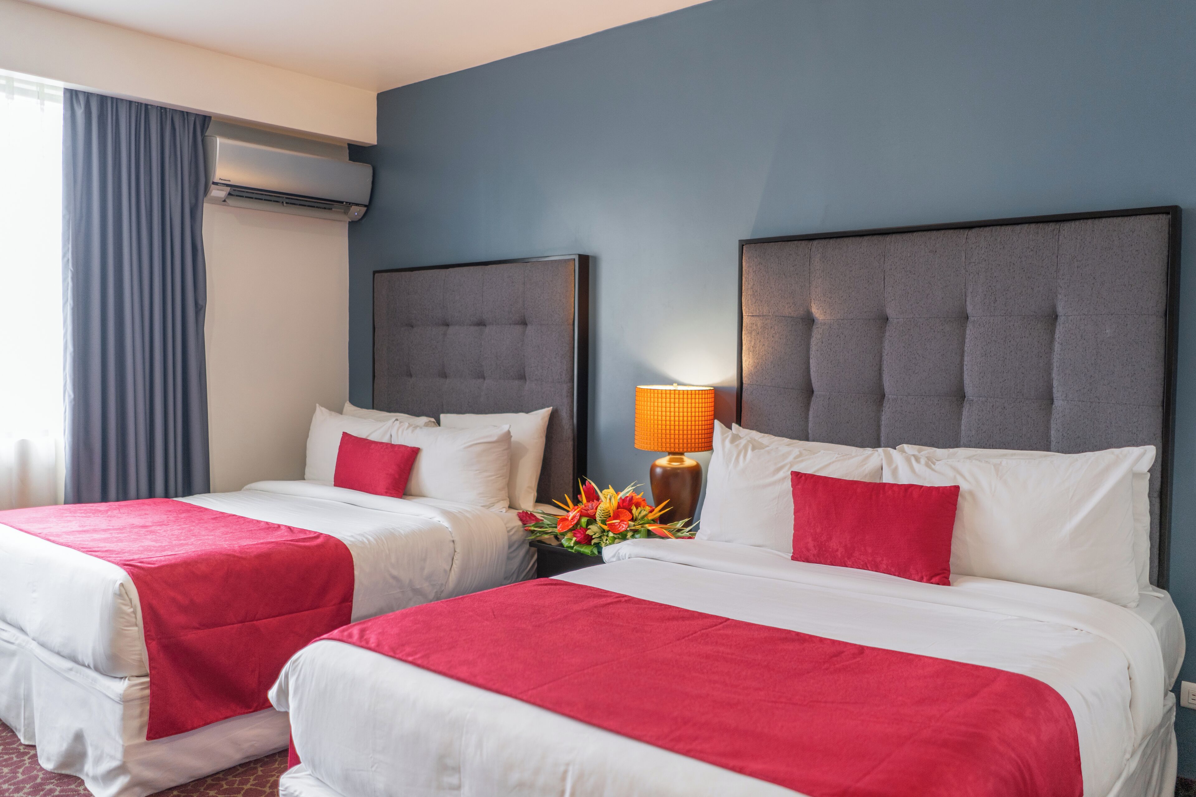 Habitación de hotel con dos camas, cada una con colcha roja y almohadas blancas, una mesita de noche con lámpara y un equipo de aire acondicionado en la pared.