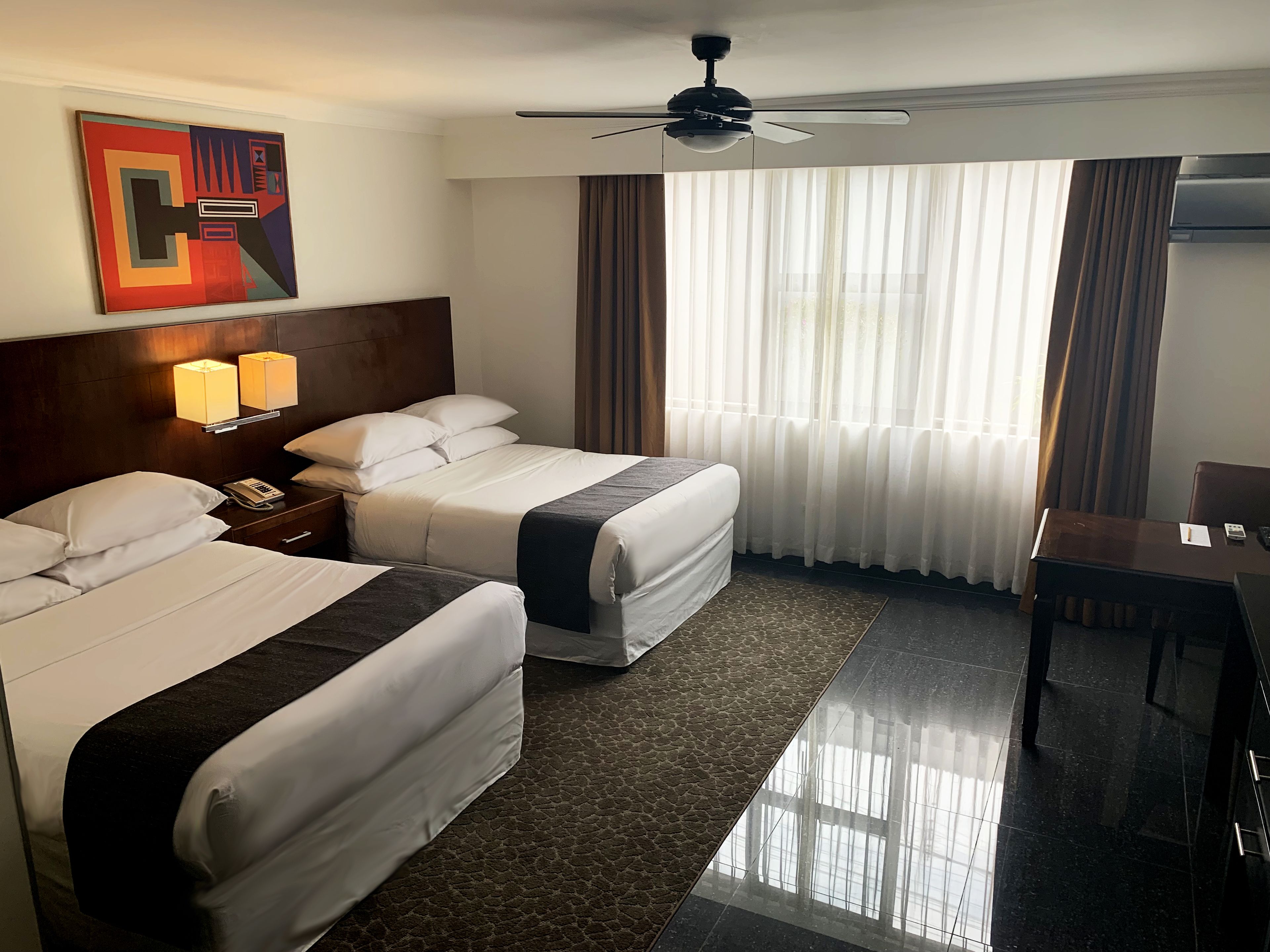 A hotel room with two beds, a desk, a ceiling fan, and a large window with curtains.