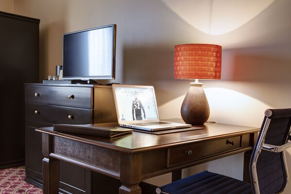 A hotel room with a wooden desk, a laptop, a television, a lamp, and a chair.