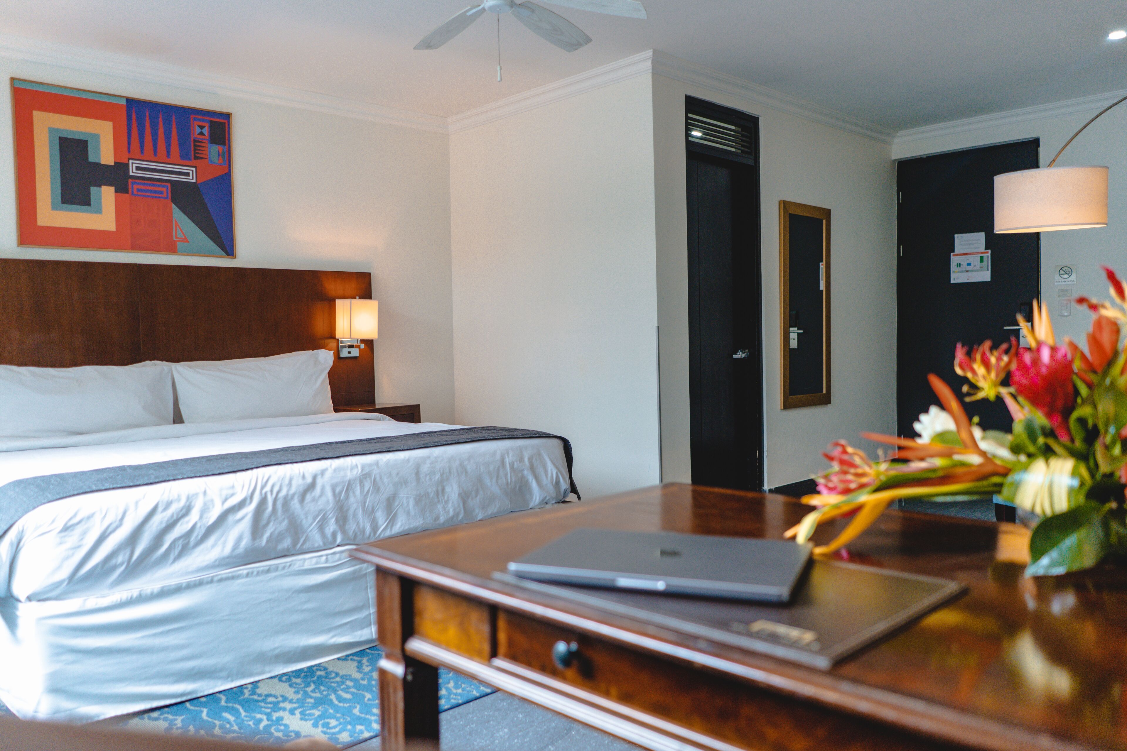 Una habitación de hotel con una cama, un escritorio con una computadora portátil, una lámpara de escritorio y un florero con flores.