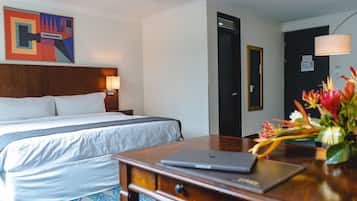 Una habitación de hotel con una cama, un escritorio con una computadora portátil, una lámpara de escritorio y un florero con flores.