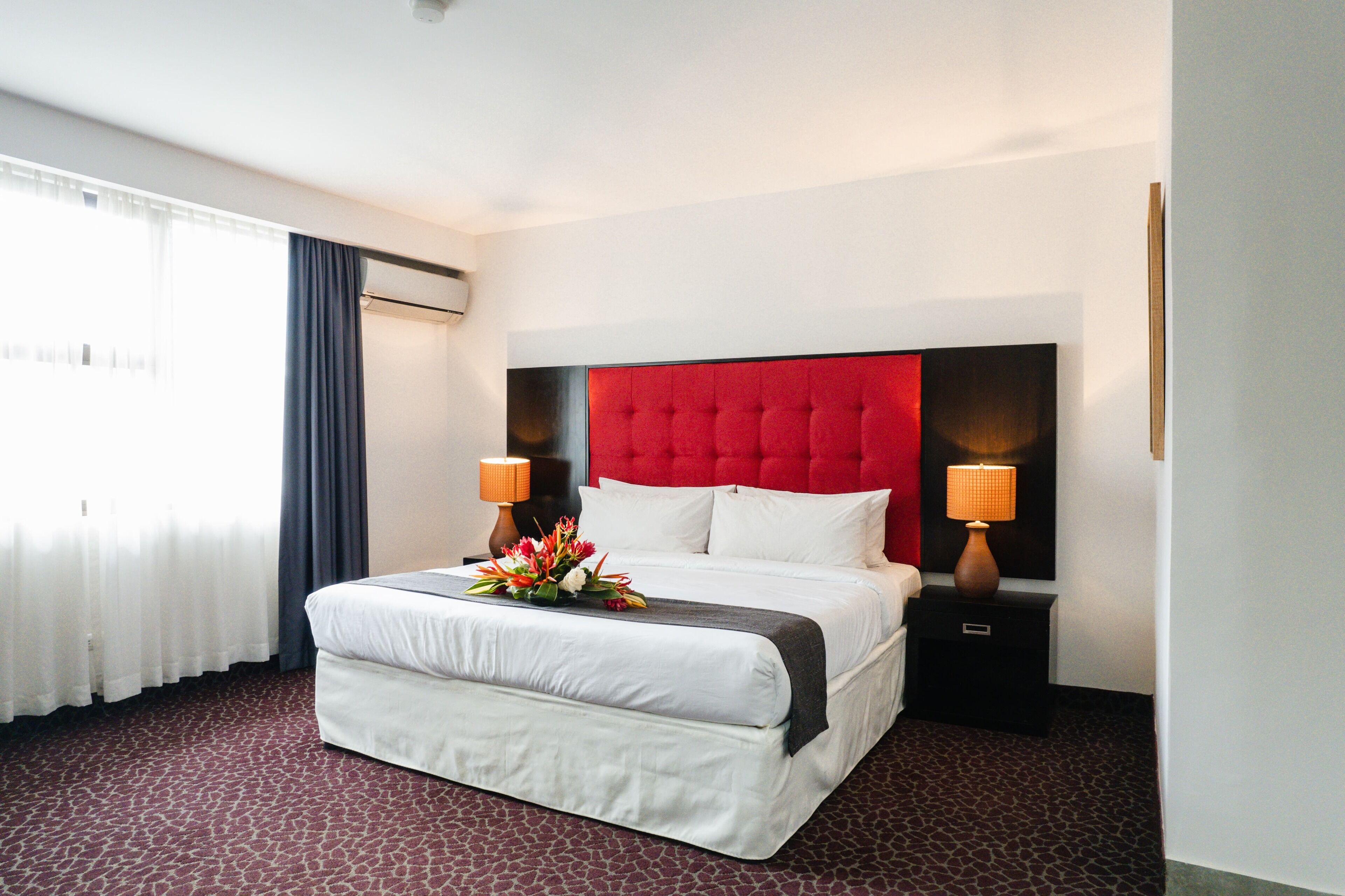 Habitación de hotel con una cama grande, dos lámparas de noche, cabecera roja y un arreglo floral sobre la cama.