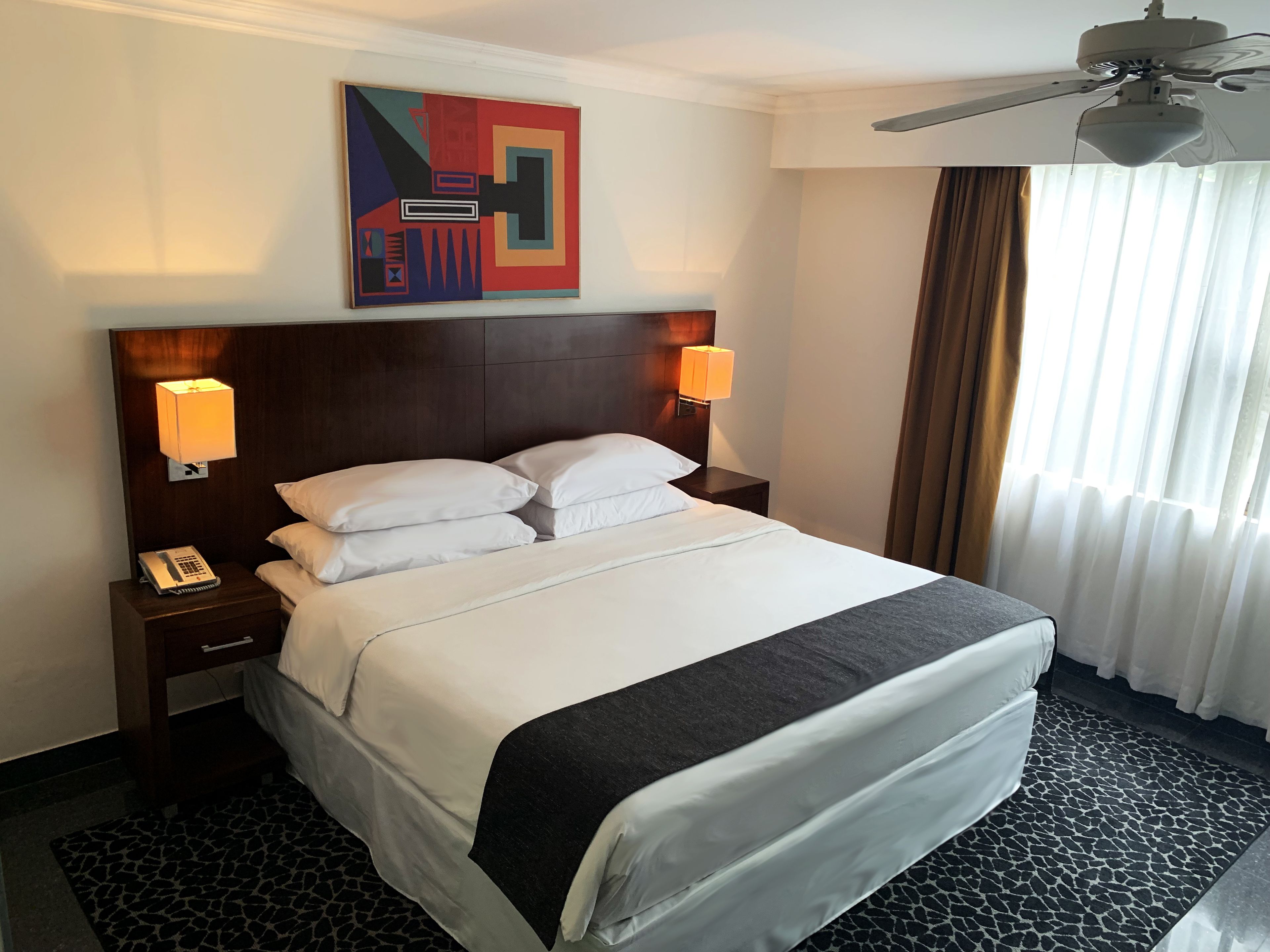 Habitación de hotel con una cama grande, dos lámparas de noche, un ventilador de techo y un cuadro abstracto moderno en la pared.