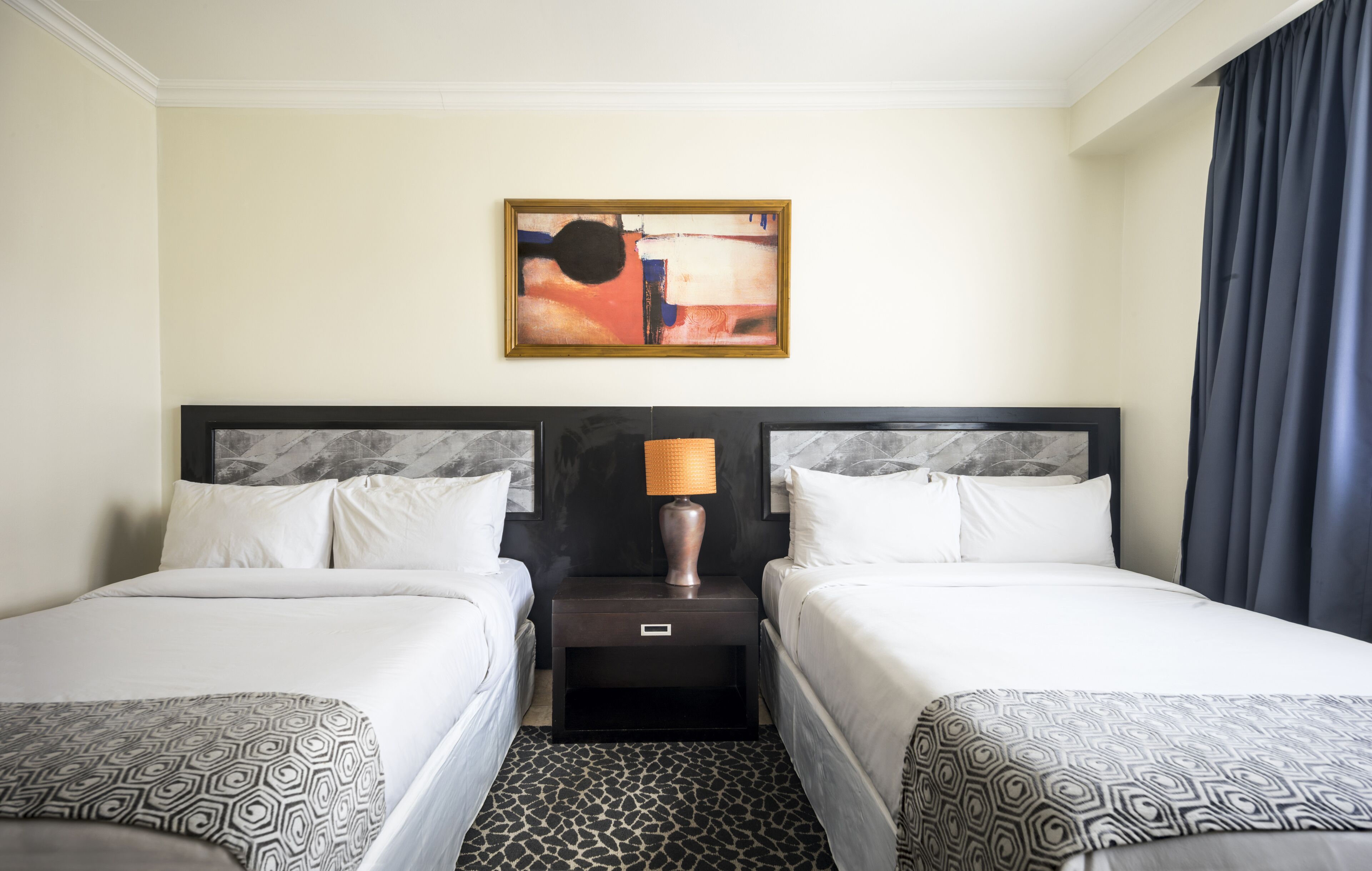 A hotel room with two beds, a nightstand with a lamp, a painting on the wall, and a patterned carpet.